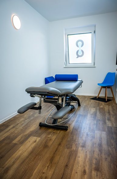 Behandlungsraum mit Massagetisch, blauer Wand und Stuhl, Holzfußboden.