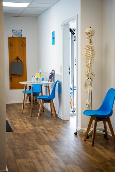 Blick in einen Flur mit blauen Stühlen, einem Tisch und einem Skelettmodell.